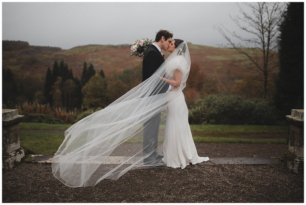  Bride and groom kissing while a long veil blows across them at their Auchen Castle wedding in Scotland. 